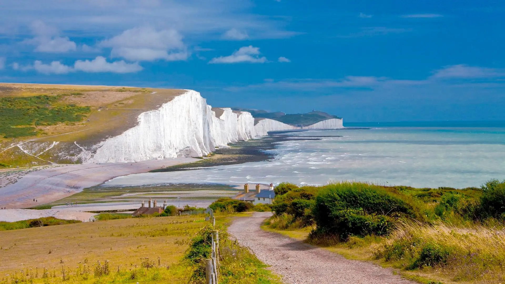 Un sentier traverse des champs côtiers vers des maisons, avec des falaises blanches, une plage et la mer bleue.